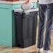 A man throwing a lid into a black rectangular Lavex Pro trash can.