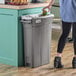 A woman opening a Lavex Pro slim rectangular gray trash can in a home kitchen.