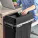 A person using a Lavex Pro black slim rectangular trash can to put a coffee cup in.