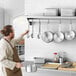 A man in an apron standing in front of a Regency stainless steel wall mounted pot rack holding a white pan and spoon.