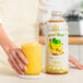 A hand holding a jar of yellow Smartfruit Sunny Banana smoothie mix next to a glass of yellow liquid.