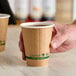 A brown compostable double wall paper hot cup with green labeling being held by a hand.
