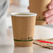 A hand holding a brown Stalk Market Planet+ paper cup with a green stripe.