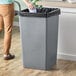 A woman putting a black liner in a Lavex gray square trash can.