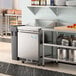 An Avantco stainless steel refrigerated sandwich prep table on a large stainless steel counter in a commercial kitchen.