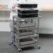 A person standing next to a Luxor gray metal serving cart with food containers on it.