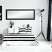 A BrandtWorks matte black mirror leaning against a wall in a white bedroom with a black and white striped bed.