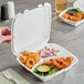 A white foam three-compartment square take out container with a hinged lid, holding fried food, curly fries, coleslaw, and a dipping sauce.