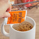 A hand holding an orange Ellis Freeze Dried Instant Decaf Coffee packet over a mug of liquid.