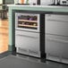 An AvaValley stainless steel wine fridge with a glass door and bottom drawer filled with wine bottles.