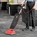 A person using a Hoover Prime Lite upright vacuum cleaner with a red cable in a corporate office cafeteria.