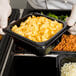 A person in a white glove holding a Cambro black plastic food pan filled with macaroni and cheese.