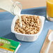 A single-serve bowl of Apple Cinnamon Cheerios cereal with milk being poured in, placed on a blue tray next to a glass of orange juice and a packaged cereal label.