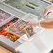 A person in gloves using tongs to serve food from a clear plastic food pan with a handle and notch in it.