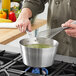 A person stirring green liquid in a Vollrath stainless steel sauce pan on a stove.