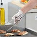 A person wearing gloves uses Choice stainless steel scalloped tongs to hold grilled food.