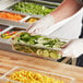 A person in gloves holding a clear polycarbonate food pan of cucumbers and corn.