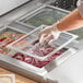 A person putting food into a clear plastic food pan using a clear polycarbonate food pan lid with a handle.