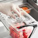 A gloved hand using a clear polycarbonate food pan lid with a notch and handle on a plastic container of tomatoes.