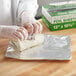 A person in gloves is using Choice interfolded pop-up foil sheets to wrap food on a table in a professional kitchen.