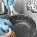 A person in blue gloves cleaning a Vigor stainless steel non-stick fry pan with a brush.