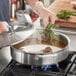 A person using a Vigor stainless steel saute pan with a lid and helper handle to cook food on a stove.