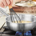 A person using a whisk to stir liquid in a Vigor Tri-Ply stainless steel saucier pan.