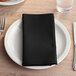 A black Choice cloth napkin folded on a white plate with silverware.