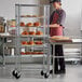 A man wearing a red apron and standing next to a Regency side load bun pan rack filled with baked goods.