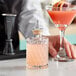 A bartender holding a Barfly glass of pink liquid next to a bottle of pink liquid.