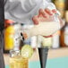 A hand using a Barfly glass bitters bottle to pour liquid into a glass with ice and a lime slice.