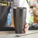 A person using a Barfly black metal cocktail shaker to pour a drink into a cup.