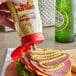A hand using Mike's Amazing Stoneground Deli Mustard to put mustard on a sandwich.