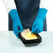 A pair of blue rubber gloves being worn by a person handling slices of cheese in a black tray.