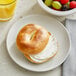 A Lender's plain bagel with cream cheese on a plate next to a bowl of fruit.