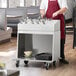 A man in a red apron standing by a ServIt flatware cart filled with utensils in a professional kitchen.