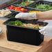A person in gloves holding a black Carlisle plastic food pan full of food.