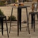 A Lancaster Table & Seating distressed gold table on a stone patio with black metal bar stools.