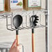 A hand holding a spoon and a spatula in a Regency chrome utensil holder on wire shelving.