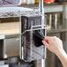 A hand putting a black plastic bag in a Regency chrome glove box holder on a wire shelf.