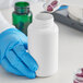 A gloved hand pouring a pink pill into a white 300cc HDPE packer bottle.