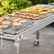 A Backyard Pro stainless steel charcoal grill with a variety of food on it in an outdoor catering setup.