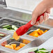 A hand holding a Carlisle red plastic salad tong over a food tray.