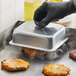 A person in black gloves using a Choice square aluminum basting cover to cook burgers on a grill.