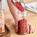 A person using a metal scoop to pour red pepper into a round plastic jar.