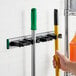 A hand using a black and white Lavex wall-mount tool holder to store a yellow broom and green mop.