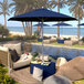 A poolside patio with California Umbrella 9' Indigo Sunbrella umbrella on a wooden base over lounge chairs.