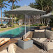 A wooden chair with a granite umbrella over it on a patio near a pool.