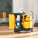 A janitor cart with shelves, a yellow vinyl zippered bag, and various cleaning supplies including a mop bucket and caution sign.
