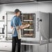 A man in a professional kitchen using a Cooking Performance Group electric combi oven to cook trays of food.